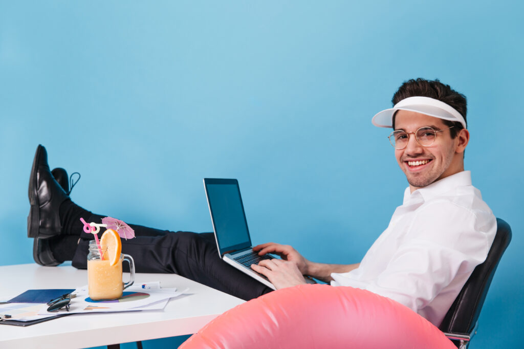 man in glasses and cap works on isolated background. guy posing with laptop, cocktail and inflatable circle.